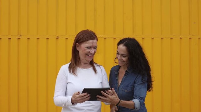 Mature woman and friend using digital tablet together against yellow background