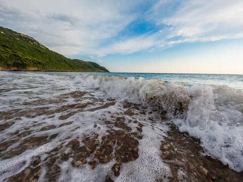 View of waves crashing on the pebble beach of the Adriatic Sea with green hills in the background under a blue sky in Montenegro.