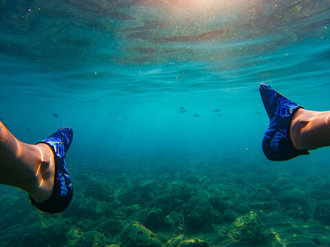 View of a person's feet wearing blue patterned water shoes while swimming in clear turquoise water over a rocky seabed in Montenegro.