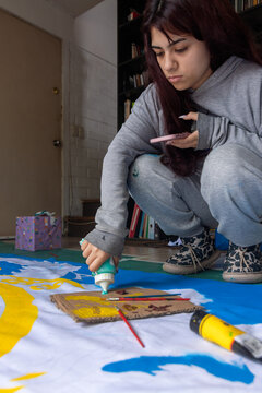 Vertical shot of young female artist squeezing paint onto palette while holding smart phone