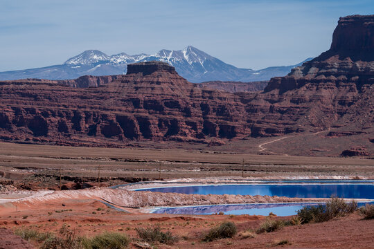 View of the vibrant blue potash evaporation ponds set against the dramatic red rock mesas and snow-capped La Sal Mountains, Moab, Utah, United States.