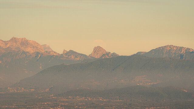 View the moonrise and the Swiss Alps illuminated by golden hour light with a hazy atmosphere