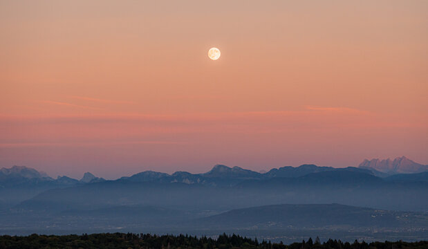 View of the full moon rising over the silhouette of the Jura mountains at dusk with soft pink and orange sky hues in Vesancy, Auvergne-Rhone-Alpes, France.
