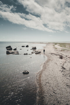 View of a person walking along a rocky beach with scattered concrete ruins in the water under a cloudy sky in Saare, Saare County, Estonia.