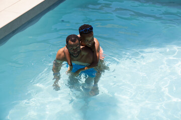 Naklejka premium African American father carrying schoolboy son playing in pool wearing blue goggles and trunks