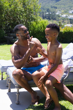 African American father and son laughing while sitting on lounge chair in swim trunks in backyard