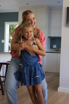Adult female in pink top hugging, laughing with child in blue dress with smartwatch in kitchen