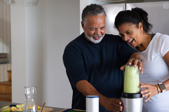 African American couple leaning and smiling using blender for green smoothie at kitchen counter