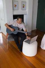 Fototapeta premium Senior Asian Chinese man sitting holding blue-gray acoustic guitar and using laptop at home