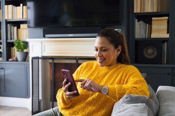 Fototapeta premium Mature African American woman in yellow sweater sitting at home on gray sofa tapping purple phone