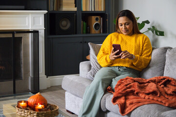 Fototapeta premium African American woman sitting right on sofa wearing mustard yellow sweater using smartphone