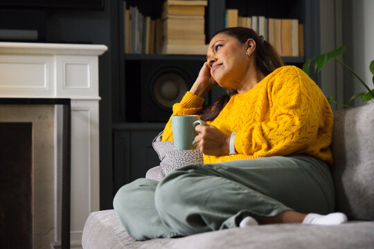 African American woman sitting on sofa in living room holding mug, in mustard sweater, copy space