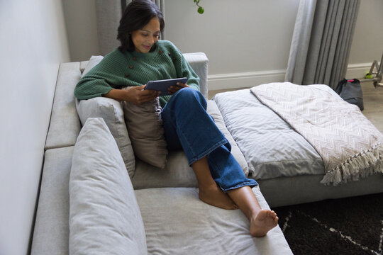 Adult African American woman reclining on sofa at home, wearing green sweater, holding tablet