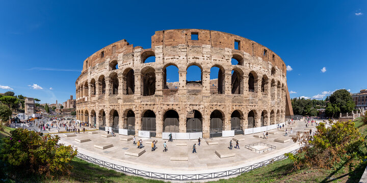 View of the Colosseum with its ancient stone arches under a clear blue sky and people walking in the plaza in Rome, Lazio, Italy.