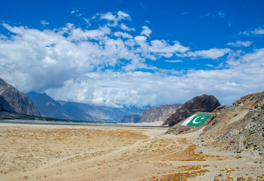 View of the arid landscape of Shigar Valley featuring a large Pakistani flag displayed on a rocky hillside under a bright blue sky with scattered clouds in Shigar, Gilgit, Pakistan.