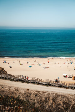 View of Praia da Bicas with wooden stairs leading to the sandy beach, colorful umbrellas, and people enjoying the sunny day by the Atlantic Ocean in Sines, Setubal, Portugal.