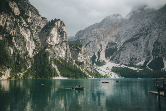 View of Pragser Wildsee with wooden rowboats on the calm water, surrounded by towering rocky mountains and pine forests under a cloudy sky in Prags, Trentino-Alto Adige, Italy.