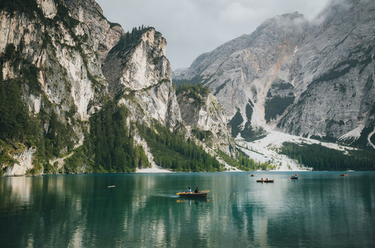 View of Pragser Wildsee with wooden rowboats on the calm turquoise water surrounded by towering limestone mountains under a cloudy sky in Prags, Trentino-Alto Adige, Italy.