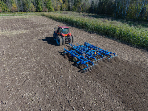Gregorville, Wisconsin, United States - 05 October 2026: Aerial view of a red tractor pulling a blue tillage implement across a harvested field with corn stalks and trees.