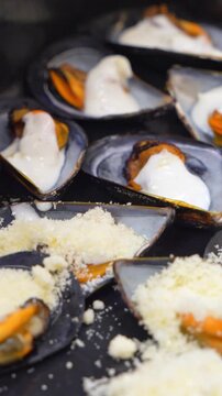 Close view of mussels on the half shell topped with creamy white sauce and a generous shower of grated cheese on a dark tray, an appetizing starter idea for a bistro menu.