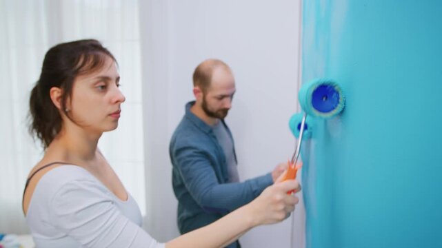 Woman uses a roller close to camera while a man paints behind her, with selective focus framing their teamwork during a practical home makeover.