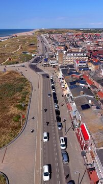 High-angle aerial drone view of Katwijk aan Zee coastal road with moving traffic, parked cars, hotels, seaside promenade, sand dunes, and North Sea coastline in South Holland on sunny summer day