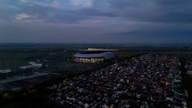 Bandung, Indonesia - 13 May 2025: Aerial view of Gelora Bandung Lautan Api Stadium illuminated at dawn, surrounded by residential rooftops and fields in Gedebage.