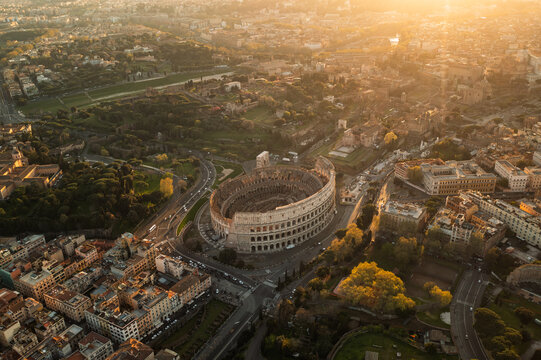 Roma, Italy - 07 April 2022: Aerial view of the Colosseum at sunset, with surrounding historic city streets, ancient ruins, and warm golden light illuminating the urban landscape.