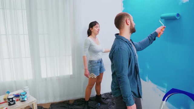 Man applies blue paint to an interior wall with a roller while a woman stands nearby, showing teamwork during an apartment makeover and DIY home improvement.