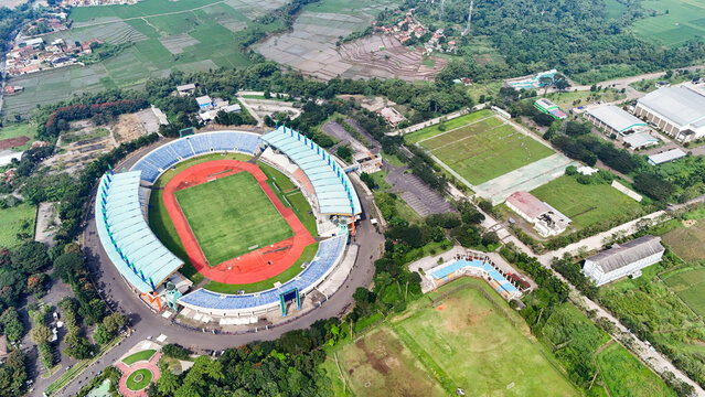Bandung, Indonesia - 12 May 2025: Aerial view of Si Jalak Harupat Stadium with its distinctive blue roof and red running track, surrounded by lush green fields and rural landscape in West Java.
