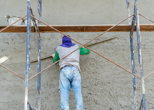 Rear view of a plasterer leveling wet mortar surface on a large concrete building wall during renovation work.