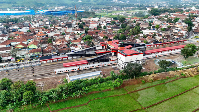 Bandung, West Java, Indonesia - 12 May 2025: Aerial view of Cicalengka Train Station with modern architecture, trains, residential rooftops, and vibrant green rice fields.