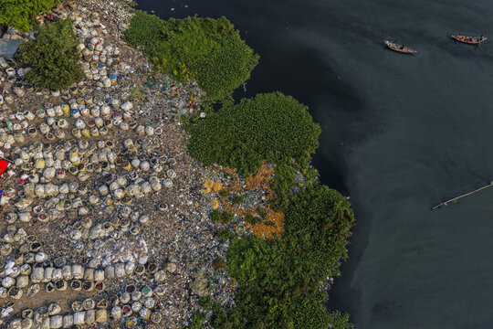 Dhaka, Dhaka Division, Bangladesh - 04 October 2026: Aerial view of Kamrangirchar showing a riverbank polluted with plastic waste and trash bags, with green vegetation and boats on the river.