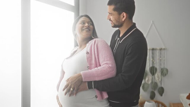 Woman and man embrace, smiling with hands on pregnant belly at window in building with macrame wall hanging visible; tender anticipation.