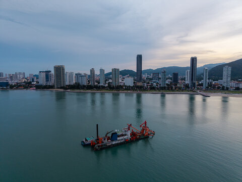 Penang, Malaysia - 01 July 2026: Aerial view of a dredging vessel operating in the sea with the city skyline and coastal buildings in the background under a cloudy sky.