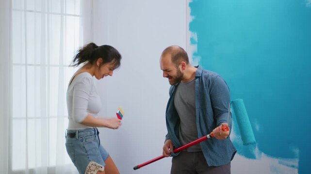 Playful couple turns painting tools into pretend microphones while redecorating a turquoise wall, adding singing, humor and joy to a hands-on DIY home makeover and shared decorating project.