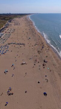 Aerial drone view of Katwijk aan Zee sandy beach packed with tourists, North Sea coastline, waves, beach bars, and coastal dunes in South Holland on sunny summer day