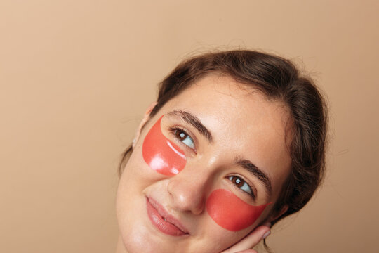 Self care, young woman with red under eye patches looks away in thoughtful three quarter pose on beige background. Concept of skincare, beauty routine and facial treatment at home.