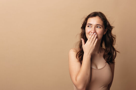 Body positivity, young woman in a beige top covers her mouth with hand and smiles, natural beauty and self acceptance concept on a beige background.