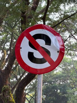 No stopping road sign with red slash over letter S against backdrop of green trees, urban traffic regulation symbol for transportation use