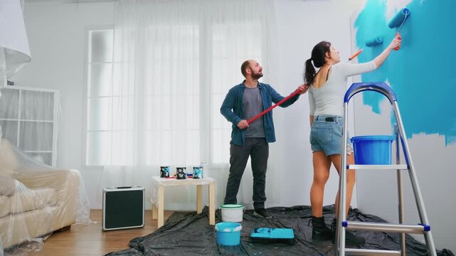 Man and woman paint a bright room blue with rollers beside a step ladder, combining teamwork, active DIY renovation and interior makeover work.