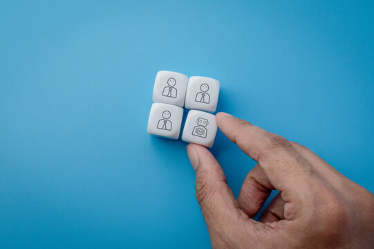 Hand placing a white cube with an AI robot icon alongside three cubes with human figures. AI workplace integration, automation, workforce transformation, job displacement, technology adoption concept.