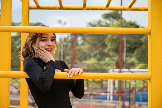 Colombian teen girl feeling surprised and happy, smiling widely, looking at the camera, posing behind yellow monkey bars at an outdoor playground