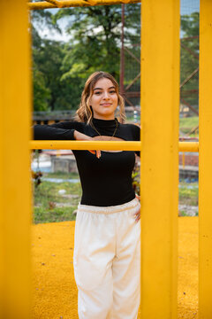 Colombian teen girl posing on a playground, wearing sportswear and smiling while leaning on yellow monkey bars, representing youth, active lifestyle, and modern street fashion
