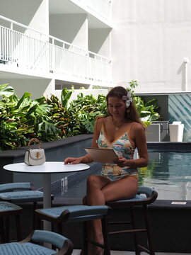 Woman in a floral swimsuit sitting at a poolside table surrounded by tropical plants at a hotel