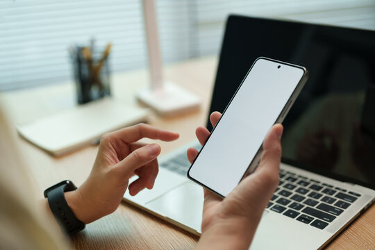 Person holds a smartphone with a blank screen beside a laptop at a desk, highlighting mobile technology, digital interaction and a clean device mockup.