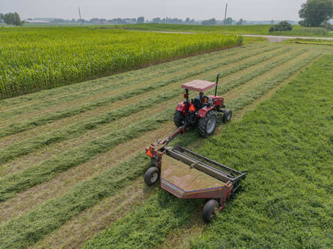 Luxemburg, Wisconsin, United States - 19 July 2026: Aerial view of a red tractor cutting hay in a lush green field with rows of mown grass and a cornfield in the background.