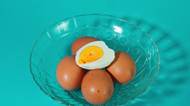 Halved hard-boiled egg resting on top of whole brown eggs in glass bowl on turquoise background