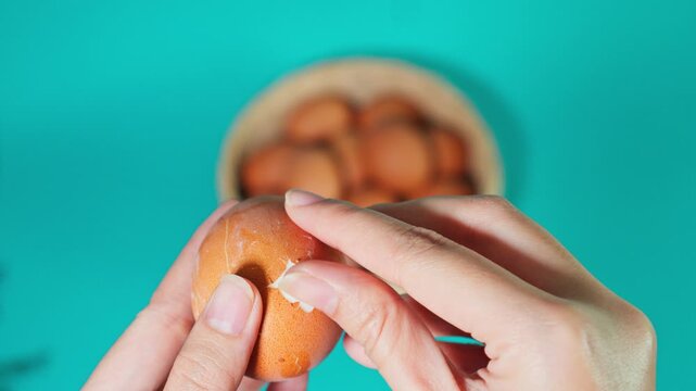 Hands peeling shell off boiled brown egg with basket of eggs in background