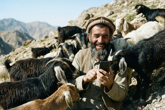 A laughing shepherd using a smartphone among a herd of goats in the mountains
